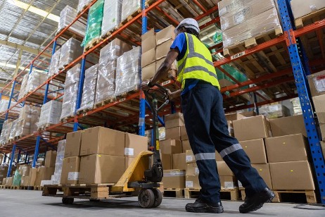 A low-angle view shows a male warehouse worker wearing a hard hat and a high-visibility vest. He is using a manual pallet jack to move a stack of boxes down an aisle. The tall warehouse racking system is visible on both sides, which is the focus of a blog about a Warehouse Racking System Safety Checklist.